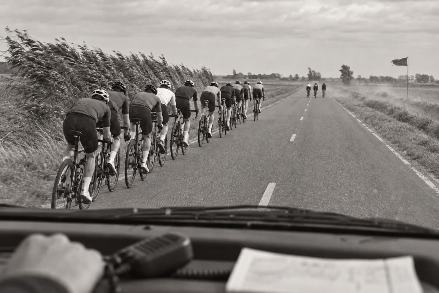 A long single-file line of riders on an exposed flat road, vegetation bent sideways by the wind